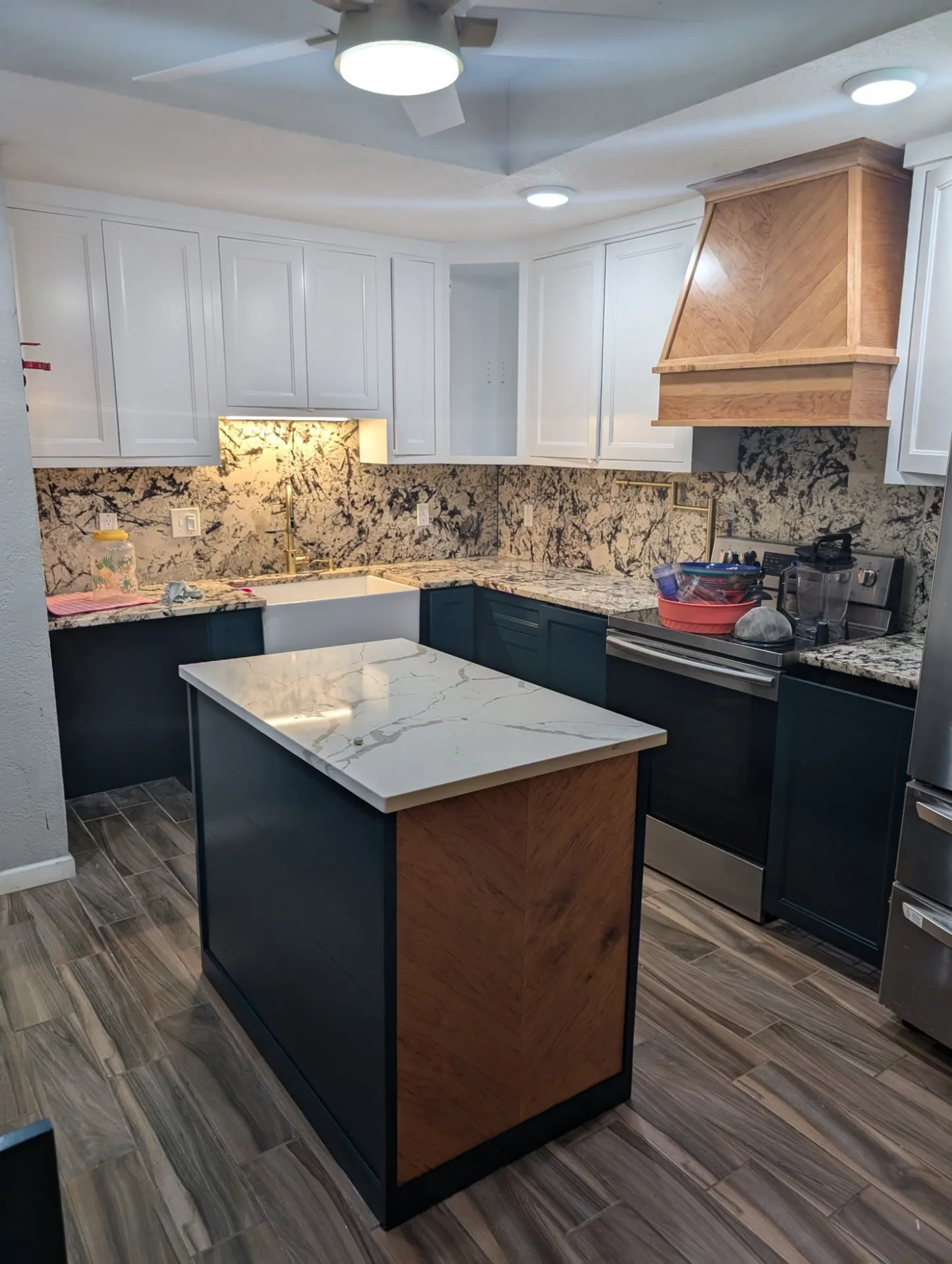 Kitchen with white upper cabinets, dark navy lower cabinets, a wood-tone range hood and a marble-look island