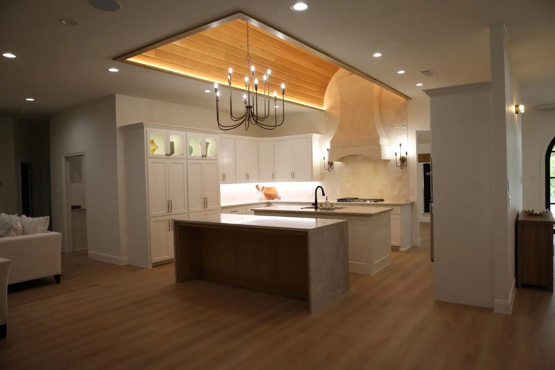 Kitchen with white cabinets, a waterfall-edge island and a lit plank-style ceiling panel with a black chandelier